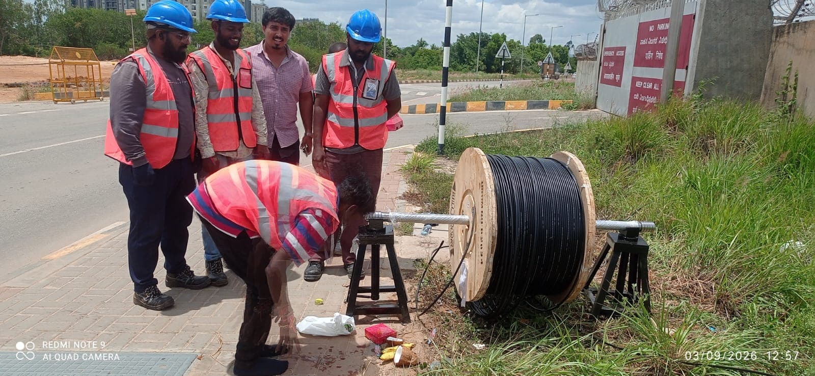A crew member guiding a reel of yellow rodder along a paved roadside route while a second crew member works at a chamber